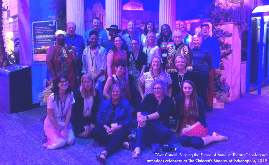 Group of 23 people gathered in a group in four rows, with the front two sitting on the floor of in a pink and purple lit space. Text on image reads "Get Crititcal: Forging the Future of Museum Theatre" conference attendees celerbate at The Children's Museum of Indianapolis, 2019."